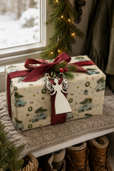 White filigree angel gift tag tied to a Badgering Home for Christmas wrapped present beside a snowy window.