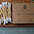 Bamboo Cotton swabs on a wooden tray with a cardboard box labeled 'New Living Cotton Swabs'.