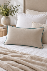 A linen cushion with duck egg piping  styled on a bed with white and beige pillows, a textured beige blanket, and a wooden side table with a vase of greenery.