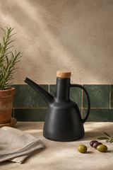 A matte black ceramic oil bottle with a cork stopper, styled on a beige stone surface with a green tiled backsplash, a potted rosemary plant, a linen napkin, and olives.