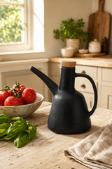 A matte black ceramic oil bottle with a cork stopper and curved handle sits on a rustic wooden table in a bright kitchen, styled with tomatoes, basil, and a linen cloth.