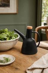 A matte black ceramic oil bottle with a cork stopper and curved handle, styled on a wooden table next to a bowl of salad in a home interior setting.
