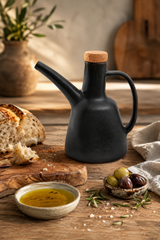Matte black ceramic oil bottle with a cork stopper, styled on a rustic wooden table with bread, olive oil, olives, and rosemary.