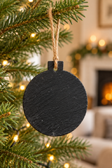 A round black slate Christmas ornament hanging from a natural jute twine string on a decorated evergreen tree, with a warm, blurred living room background featuring a fireplace.
