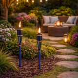 Two blue garden torches with flames in a garden setting with flowers and seating area.