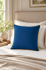 A royal blue square throw pillow with a thin golden yellow trim, styled on a bed with white pillows and a beige headboard in a bright bedroom setting.