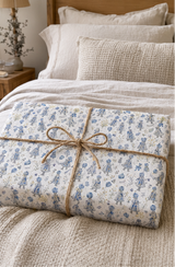 A wrapped rectangular Gift tied with twine sits on a bed with cream bedding and pillows. The package has a white background with a delicate blue girls in dress with blue bonnet pattern.