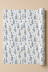 A roll of wrapping paper unrolled to show a repeating pattern of blue and gray Illustrated Water Colour girls in dresses on a white background, set against a solid beige backdrop.