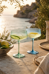 Two cocktail glasses with a lemon garnish on a table by a body of water. Blue & Green Twist Champagne Coupe Glasses – Set of 2