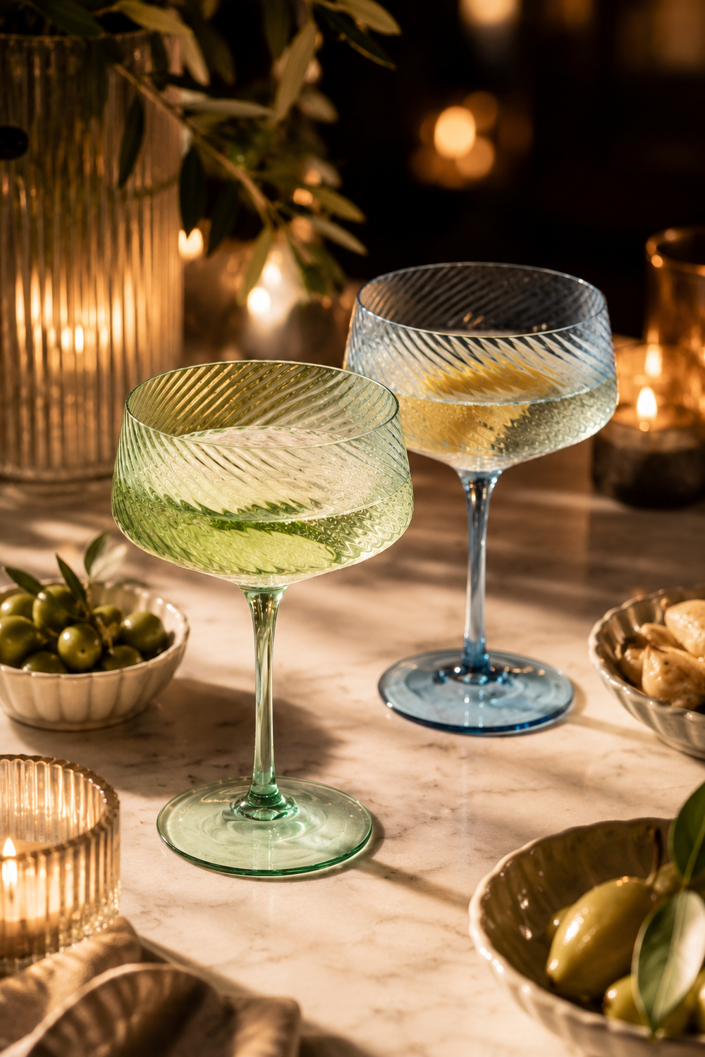 Blue & Green Twist Champagne Coupe Glasses – Set of 2  on a wooden table with candles and snacks in the background