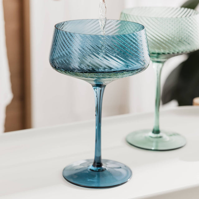 Blue glass champagne coupe glass on a white surface with a blurred background