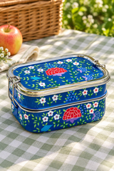 A blue metal bento lunch box with a floral and mushroom pattern, set on a green and white checkered picnic blanket outdoors with a basket and apple in the background.