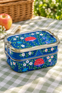 A blue metal bento lunch box with a floral and mushroom pattern, set on a green and white checkered picnic blanket outdoors with a basket and apple in the background.
