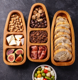 Wooden appetizer tray with sections of nuts, figs, bread, and other snacks on a dark surface.