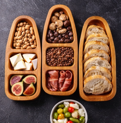 Wooden appetizer tray with sections of nuts, figs, bread, and other snacks on a dark surface.