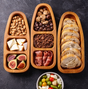 Wooden appetizer tray with sections of nuts, figs, bread, and other snacks on a dark surface.