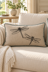 A rectangular linen cushion with black dragonfly print and a duck egg piped border, resting on a cream-colored sofa in a bright, modern living room.