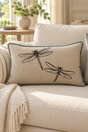 A rectangular linen cushion with black dragonfly print and a duck egg piped border, resting on a cream-colored sofa in a bright, modern living room.