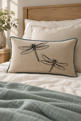 A rectangular linen cushion with black dragonfly print and a duck egg border, styled on a bed with white linens and a wooden headboard.
