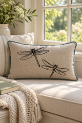 A rectangular linen throw pillow with black dragonfly printed and a duck egg border, styled on a sunlit window seat beside a cream knit blanket and a striped pillow.
