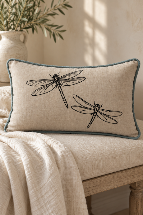 A rectangular linen throw pillow with painted dragonflies and a duck egg border, resting on a light-colored wooden bench with a neutral background.