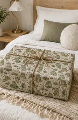 A rectangular gift box wrapped in cream-coloured wrapping paper with a green Frog with books and nature print, tied with a natural jute twine bow, resting on a cozy bed with white linens and a wooden headboard.