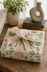 A gift box wrapped in cream wrapping paper with Frog with books pattern and tied with a natural burlap ribbon, placed on a wooden table with a potted plant and ceramic vase in the background.
