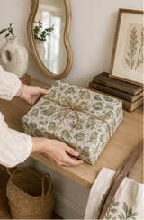 A person's hands are shown holding a wrapped gift with a green Frogs with books patterned paper and a simple brown twine bow, set on a wooden table in a cozy, styled room.