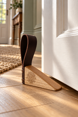 A wooden and leather door stopper resting on a light wood floor next to a white door, with a cozy interior room visible in the background.