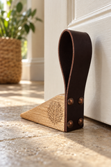 A leather and wood door stopper resting on a stone floor in front of a white door, with a blurred potted plant in the background.