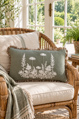 Rectangular sage botanical cushion with natural linen frill on a wicker chair beside French windows.