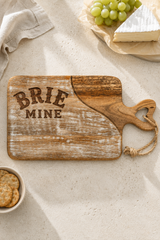 A rustic wooden serving board with a heart-shaped handle and engraved text reading 'BRIE MINE' on a light stone surface, styled with brie cheese, grapes, crackers, and a cloth napkin.