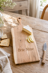 A personalized wooden cheese board with the engraved text 'Brie Mine' on a rustic wooden table, styled with a wedge of brie cheese, sliced brie, crackers, a knife, and a napkin.
