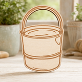 A laser-cut wooden bucket ornament or plaque with a natural light wood finish and darker brown engraved lines, standing on a light wooden surface with a blurred rustic background.