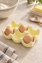 A pale yellow ceramic egg carton holding four brown eggs, styled on a light stone countertop with baking ingredients and utensils in the background.