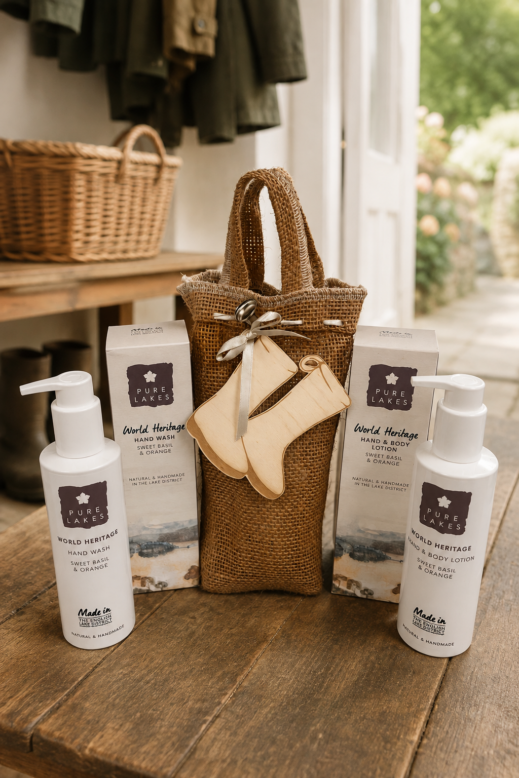 A gift set from World Heritage Handmade on a rustic wooden table, featuring two white pump bottles, two white product boxes, a brown burlap tote bag, and a small beige leather boot-shaped pouch tied with a ribbon.