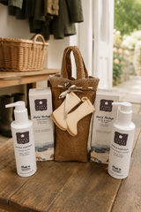 A gift set from World Heritage Handmade on a rustic wooden table, featuring two white pump bottles, two white product boxes, a brown burlap tote bag, and a small beige leather boot-shaped pouch tied with a ribbon.