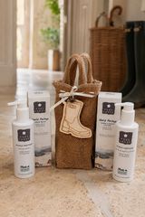 A gift set of World Heritage Cleanse products arranged on a stone surface, including two white pump bottles, two white boxes, a burlap tote bag, and a wooden boot-shaped tag, with a softly blurred indoor background.