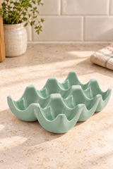 A sage green ceramic egg-shaped dish with six compartments, styled on a beige stone countertop with a tiled backsplash and a potted plant in the background.