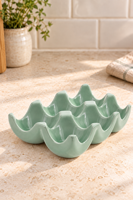 A sage green ceramic egg-shaped dish with six compartments, styled on a beige stone countertop with a tiled backsplash and a potted plant in the background.
