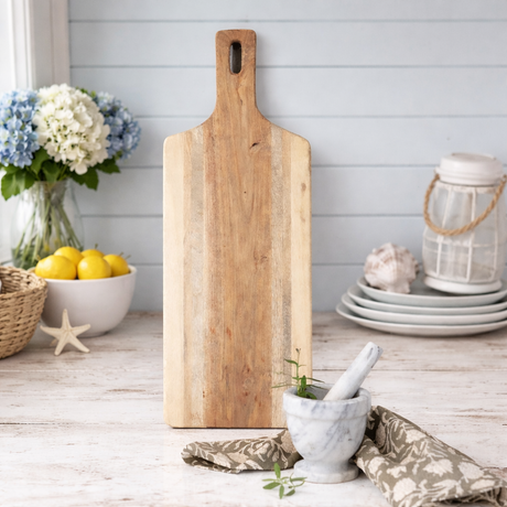 Wooden cutting board on a wooden surface with lemons, flowers, and kitchen items.