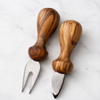 Set of wooden cheese knives with a rustic setting featuring cheese and olives.