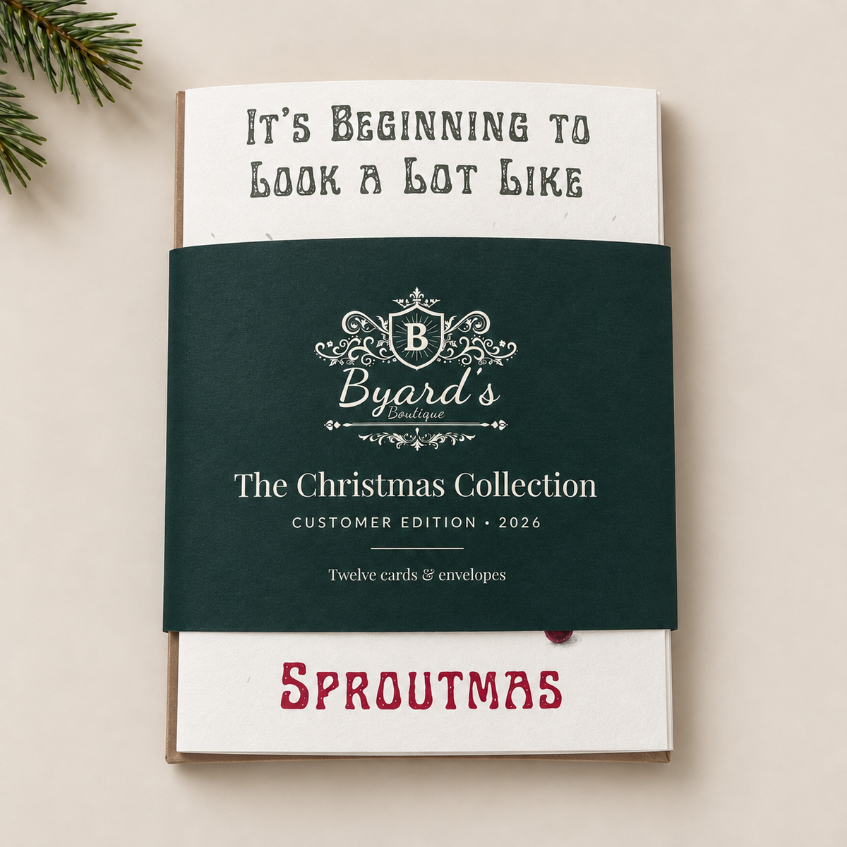 A card & envelope collection wrapped with a dark green paper band reading 'Byard's The Christmas Collection Customer Edition 2026.' The top card says 'It's Beginning to Look a Lot Like' and the bottom of the card says 'SPROUTMAS.