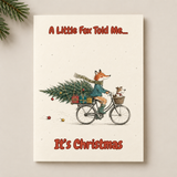 A rectangular Christmas greeting card or print with an illustrated fox riding a bicycle with a Christmas tree and presents, bordered by pine branches at the top left. The text reads “A Little Fox Told Me.