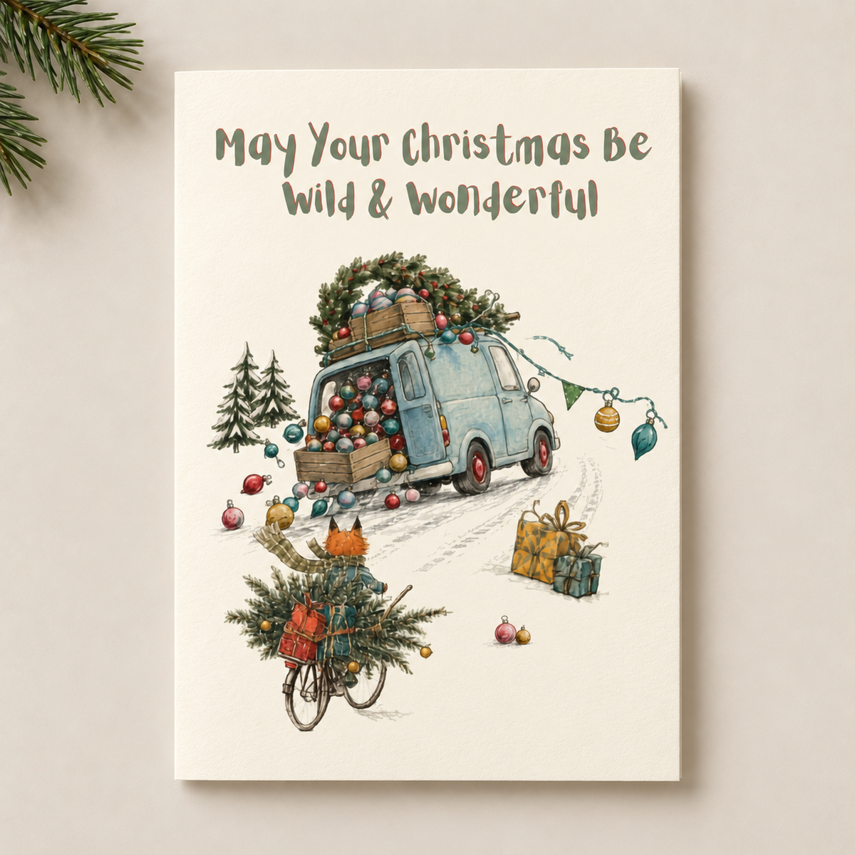 A vertical Christmas greeting card with the text 'May Your Christmas Be Wild & Wonderful' at the top, featuring an illustration of a light blue van loaded with Christmas trees and ornaments, with a bicycle carrying a Christmas tree in the foreground. The card is photographed standing upright against a light beige background with pine branches in the top-left corner.