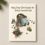 A vertical Christmas greeting card with the text 'May Your Christmas Be Wild & Wonderful' at the top, featuring an illustration of a light blue van loaded with Christmas trees and ornaments, with a bicycle carrying a Christmas tree in the foreground. The card is photographed standing upright against a light beige background with pine branches in the top-left corner.
