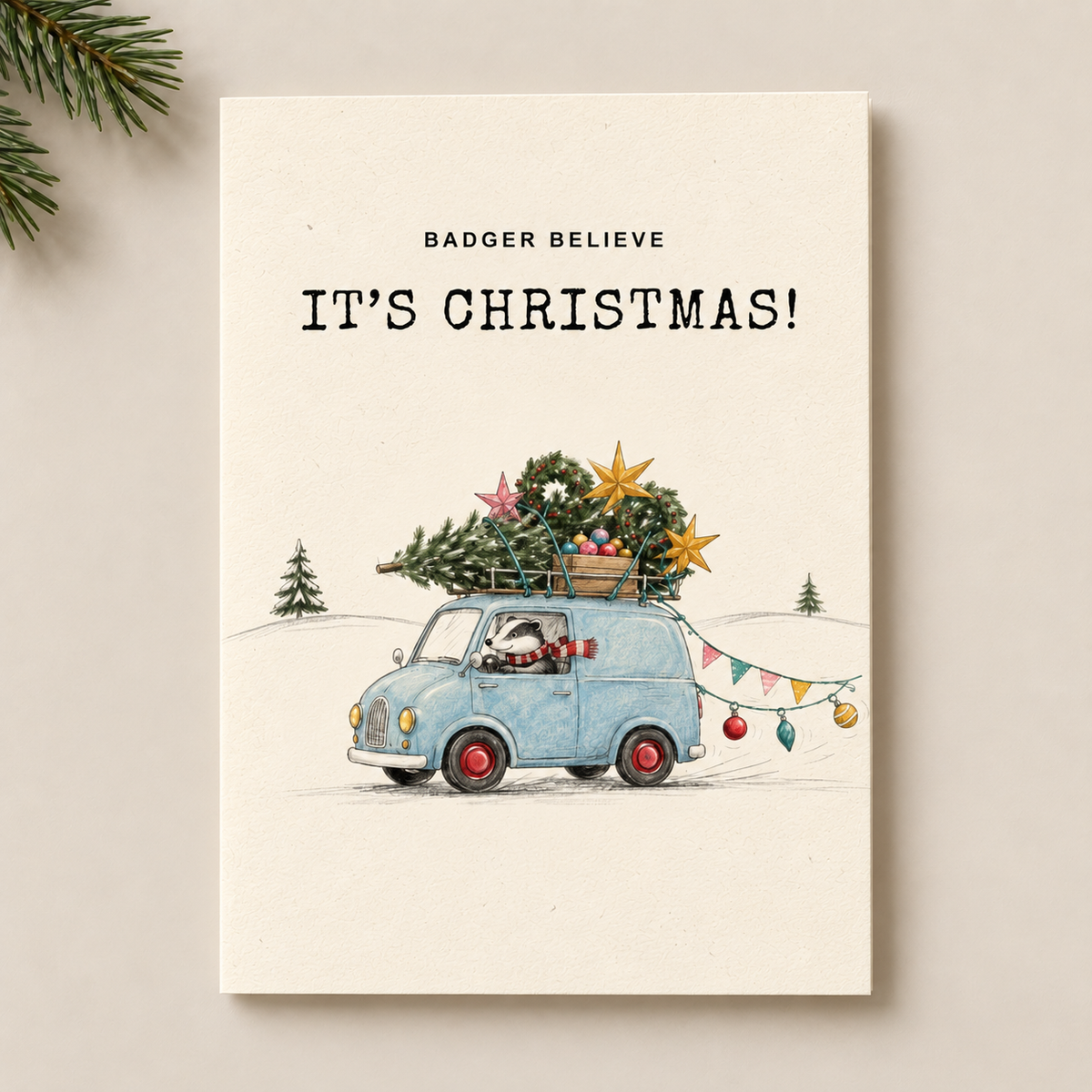 A square greeting card with a cream-colored background featuring a watercolor illustration of a light blue vintage car carrying a Christmas tree on its roof. The text 'BADGER BELIEVE' and 'IT'S CHRISTMAS!' are printed at the top.