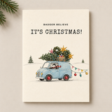 A square greeting card with a cream-colored background featuring a watercolor illustration of a light blue vintage car carrying a Christmas tree on its roof. The text 'BADGER BELIEVE' and 'IT'S CHRISTMAS!' are printed at the top.