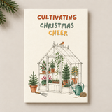 A vertical greeting card with the text 'CULTIVATING CHRISTMAS CHEER' above a hand-drawn greenhouse illustration with potted plants, a bird on the roof, and gardening tools. The card is photographed against a light neutral background with a pine branch entering from the top left.