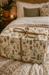 Illustrated Christmas wrapping paper featuring glasshouses, potted fir trees, holly, ivy, robins, terracotta pots, watering cans, hand forks, twine, seed trays, cyclamen and citrus trees. The pencil-sketch design uses muted green, terracotta, berry red, orange and pink on a warm ivory background.